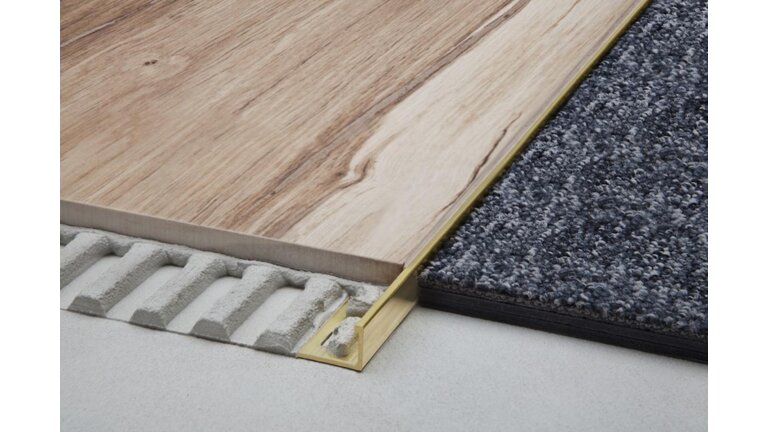 Ein Messingprofil bildet die präzise und saubere Verbindung zwischen einer Holzoptik-Laminatfläche und einem grauem Teppich.